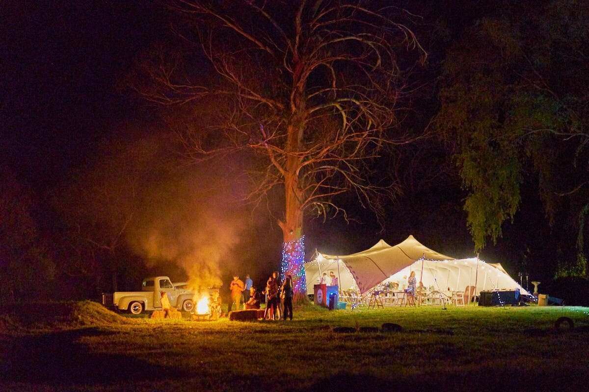 Nighttime outdoor camping scene with a bonfire, white tent, and festive lights under a large tree.