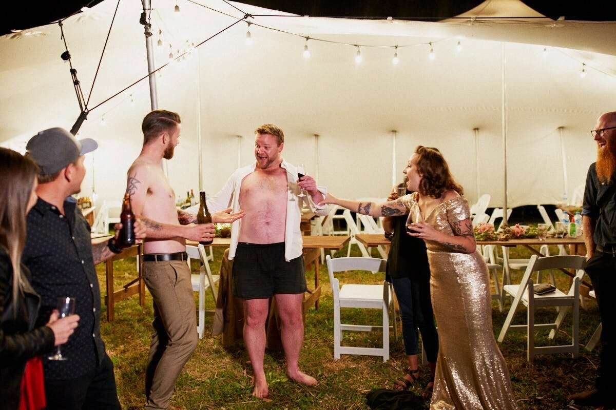 Group of people socializing at an outdoor tent event, with one man in boxer shorts holding a drink.