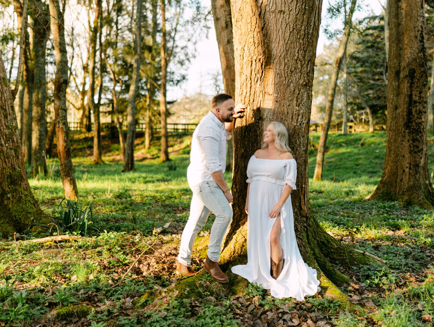 Couple in white outfits posing by a large tree in a sunlit forest during a maternity photoshoot.