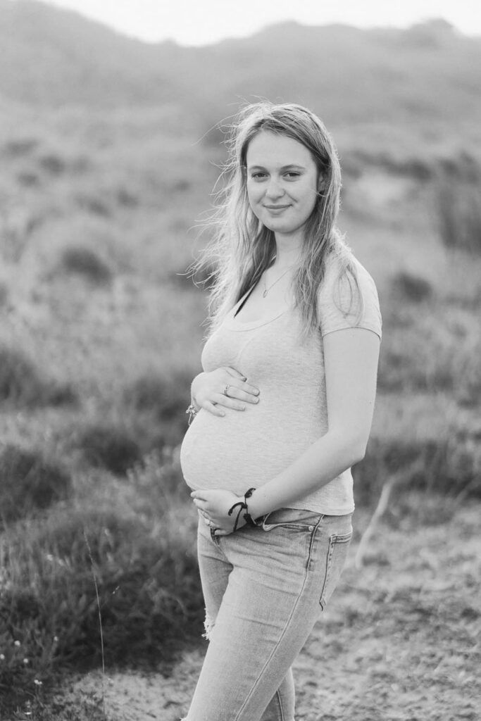 Black and white portrait of a smiling pregnant woman cradling her baby bump outdoors in a natural field setting.