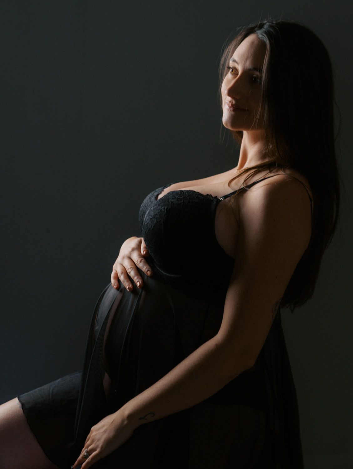 Elegant pregnant woman in black dress cradling her baby bump in a dramatic dark studio portrait.