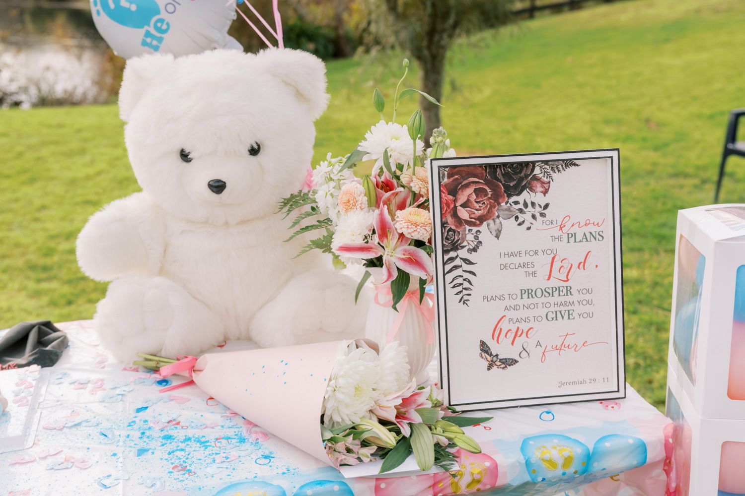 White teddy bear beside floral bouquet and inspirational sign on decorated table at outdoor baby shower event.
