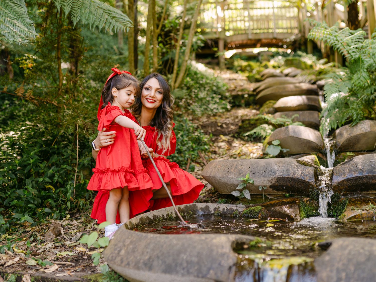 Mother and daughter in matching red dresses sitting by a garden stream surrounded by lush green ferns and rocks.