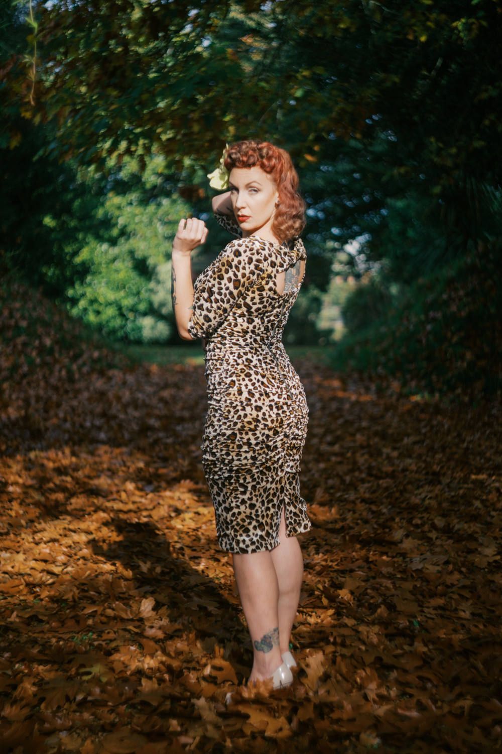Red-haired woman in leopard print dress poses among autumn leaves in a forest path.
