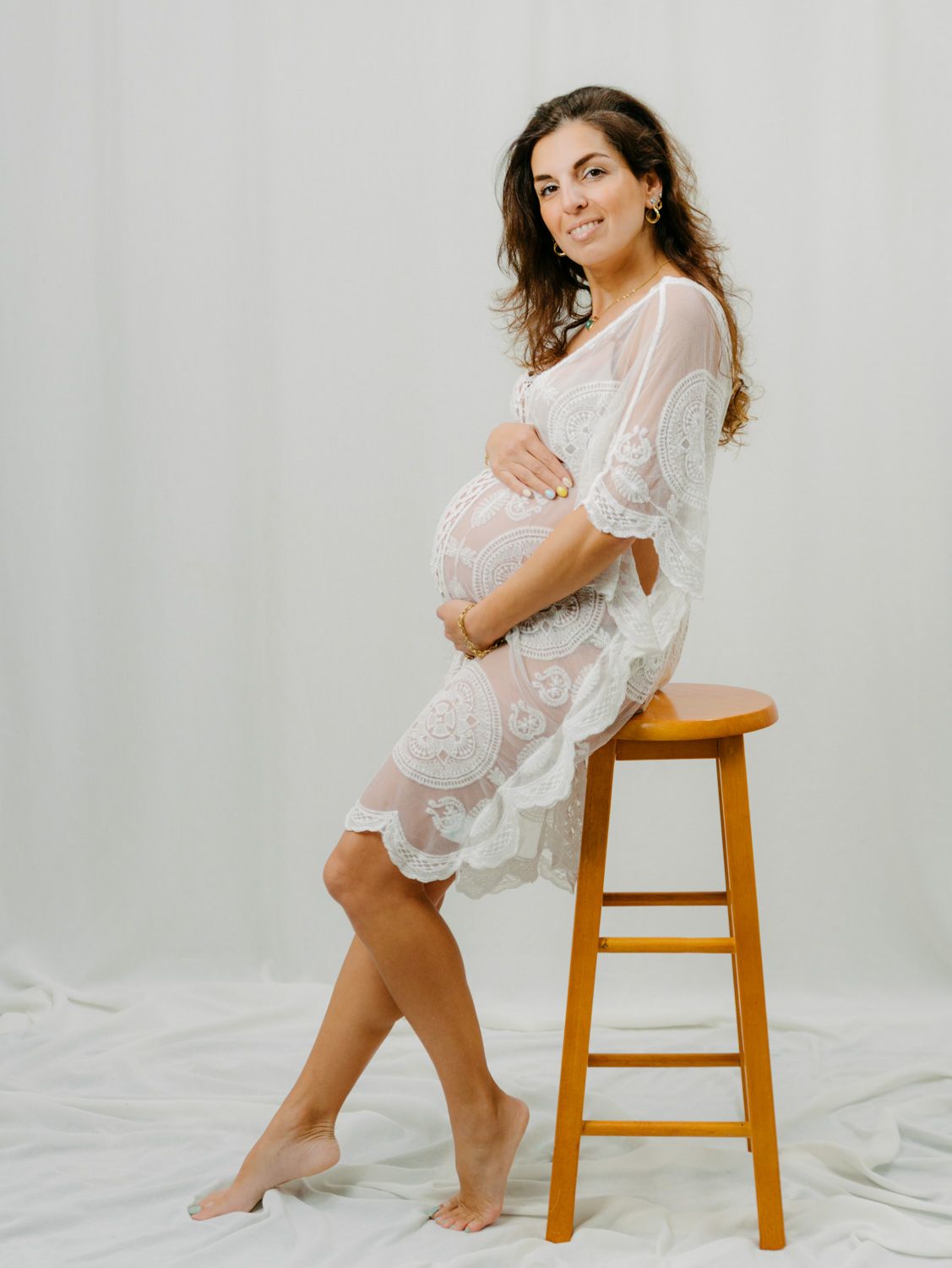 Pregnant woman in white lace dress posing on wooden stool in studio maternity photo shoot.