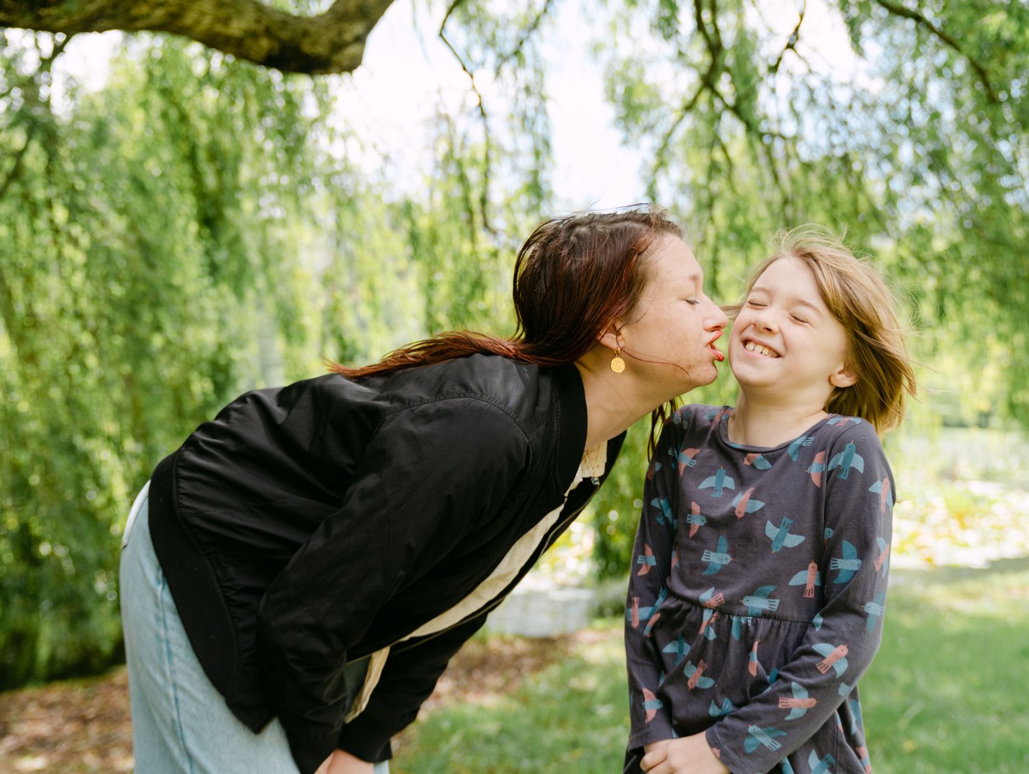A mother lovingly kisses her laughing young daughter on the cheek outdoors under a willow tree.