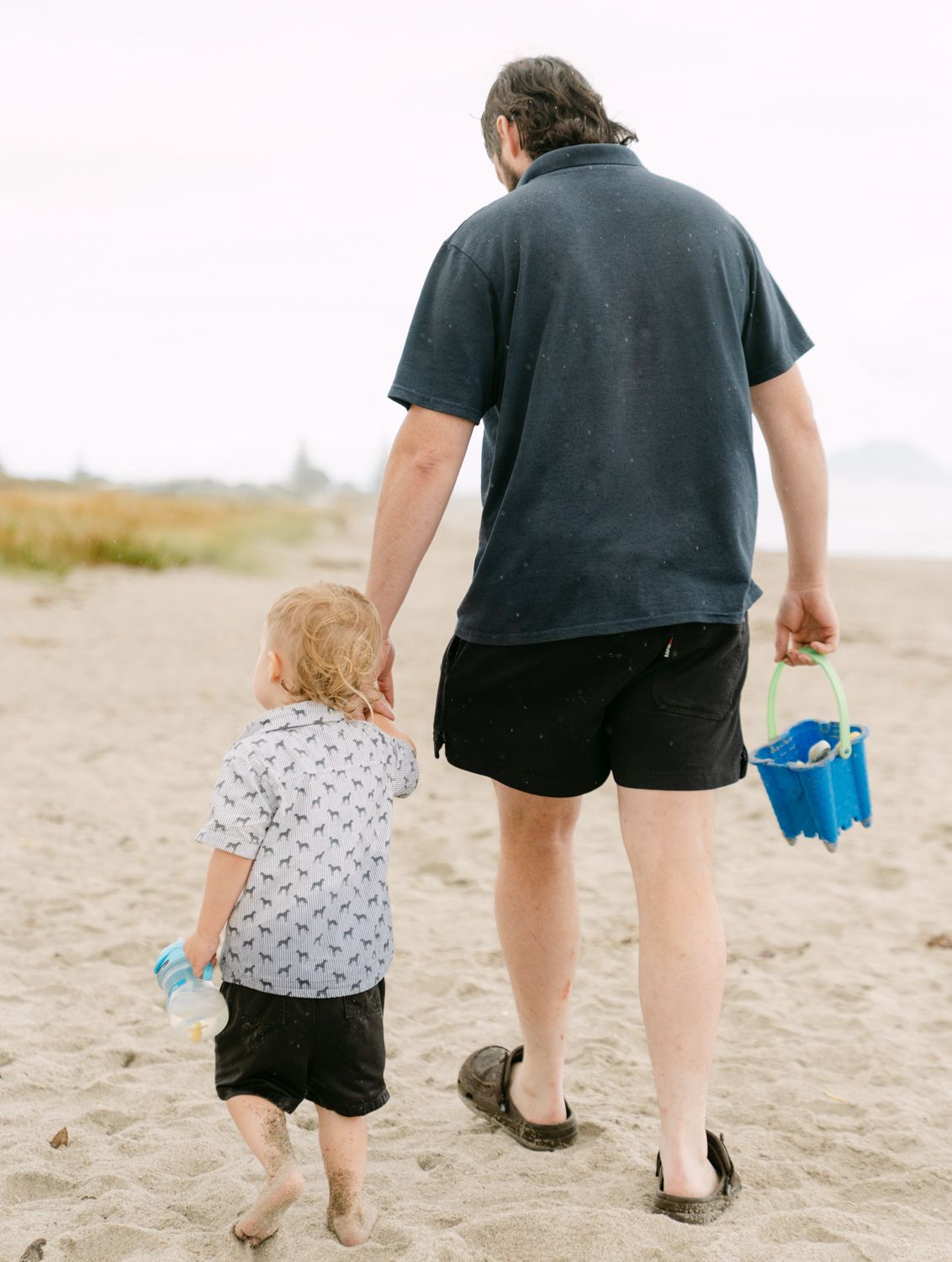 Adult and toddler walk hand-in-hand on sandy beach, man carrying blue bucket, both wearing casual summer clothing.