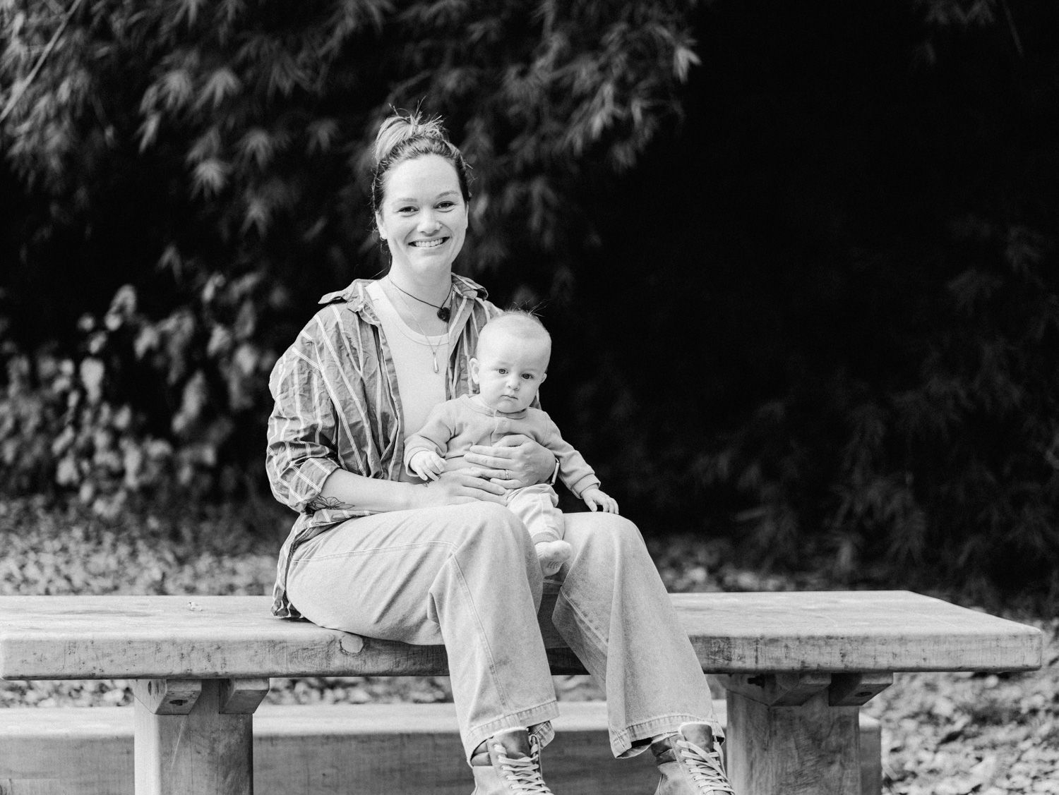 Black and white photo of a smiling woman sitting on a wooden bench outdoors, holding a baby on her lap.
