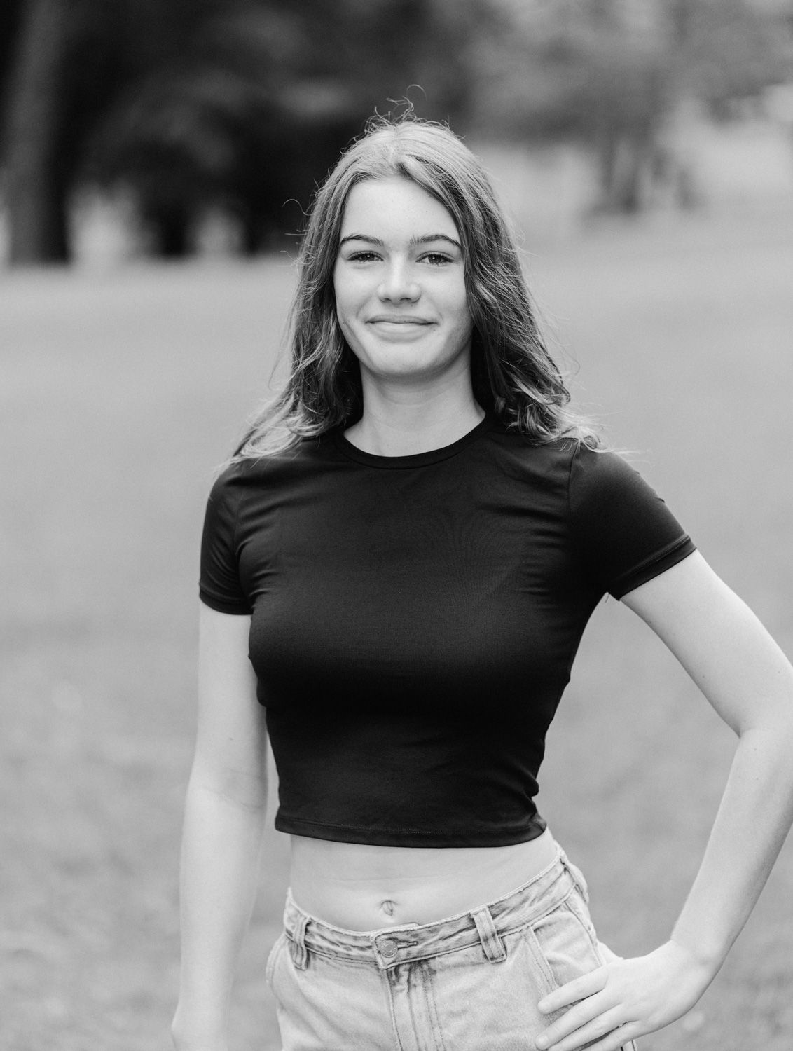 Young woman in black crop top and jeans poses outdoors in a park, smiling confidently in black and white photo.