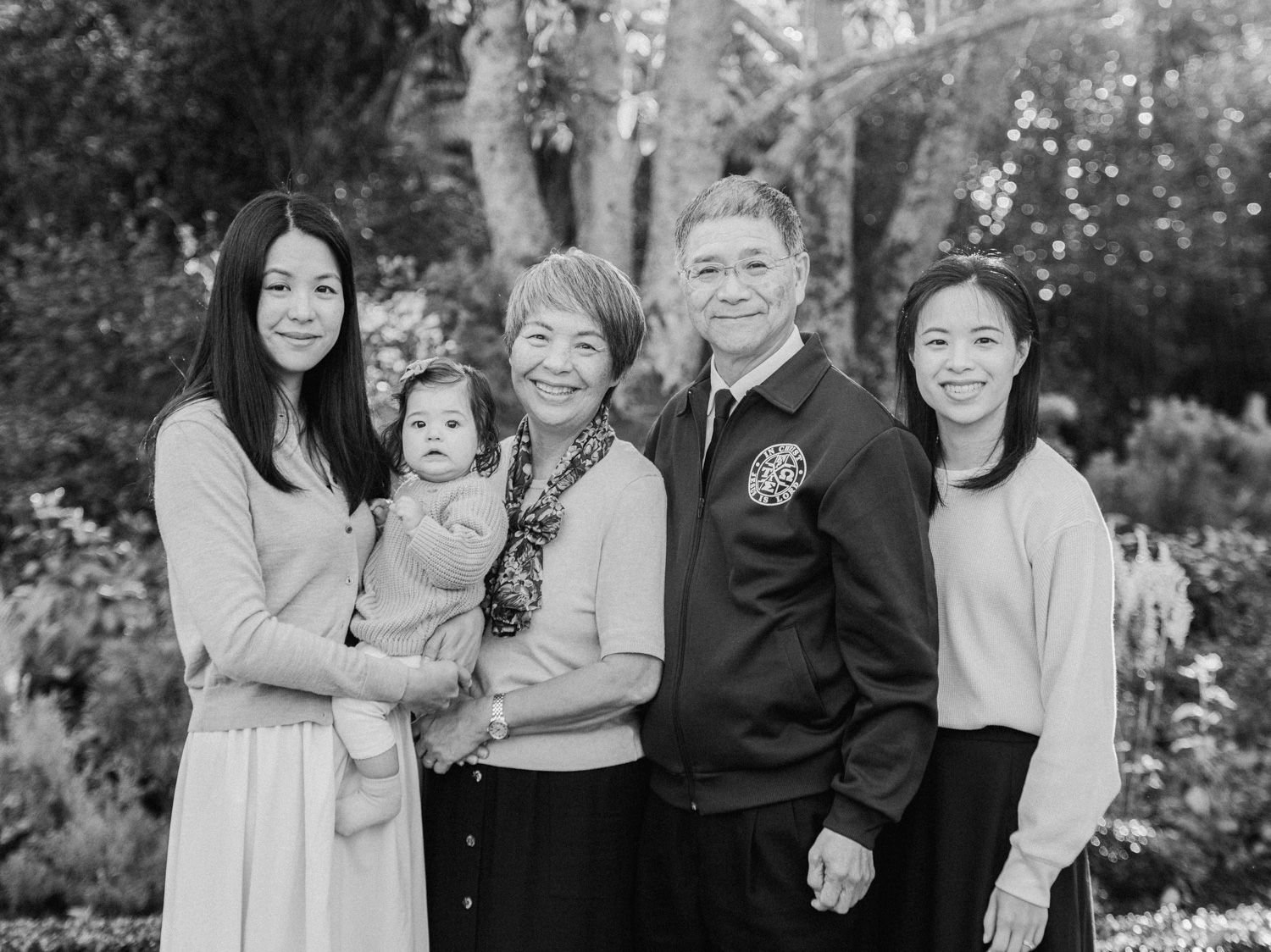 Black and white family portrait of five generations posing together in a lush garden setting.