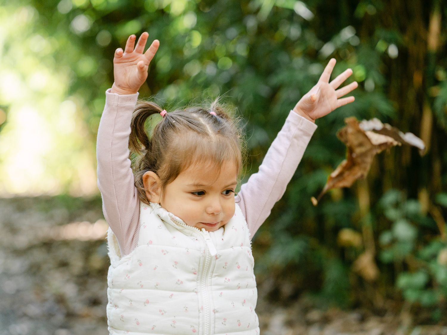 Joyful toddler girl in white vest raising arms outdoors among green trees.
