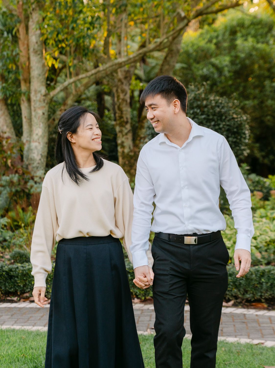 A smiling couple holds hands and walks together in a lush garden, dressed in elegant casual attire.