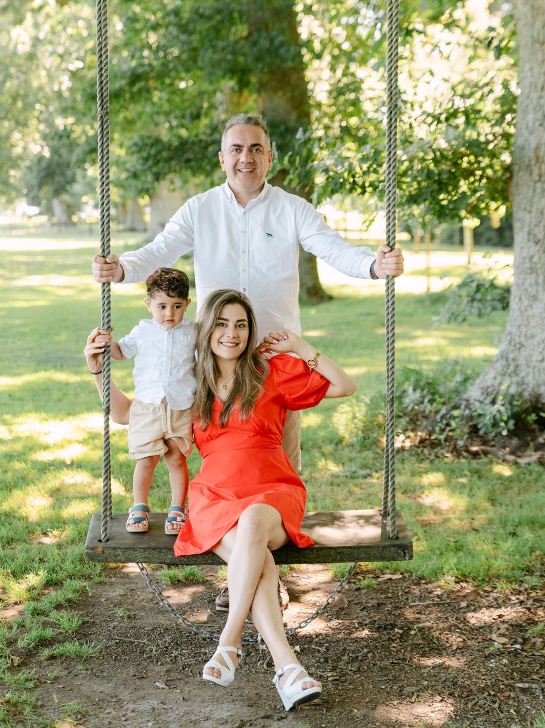 Family of three posing on a wooden rope swing in a lush green park, mother in red dress, father in white shirt.