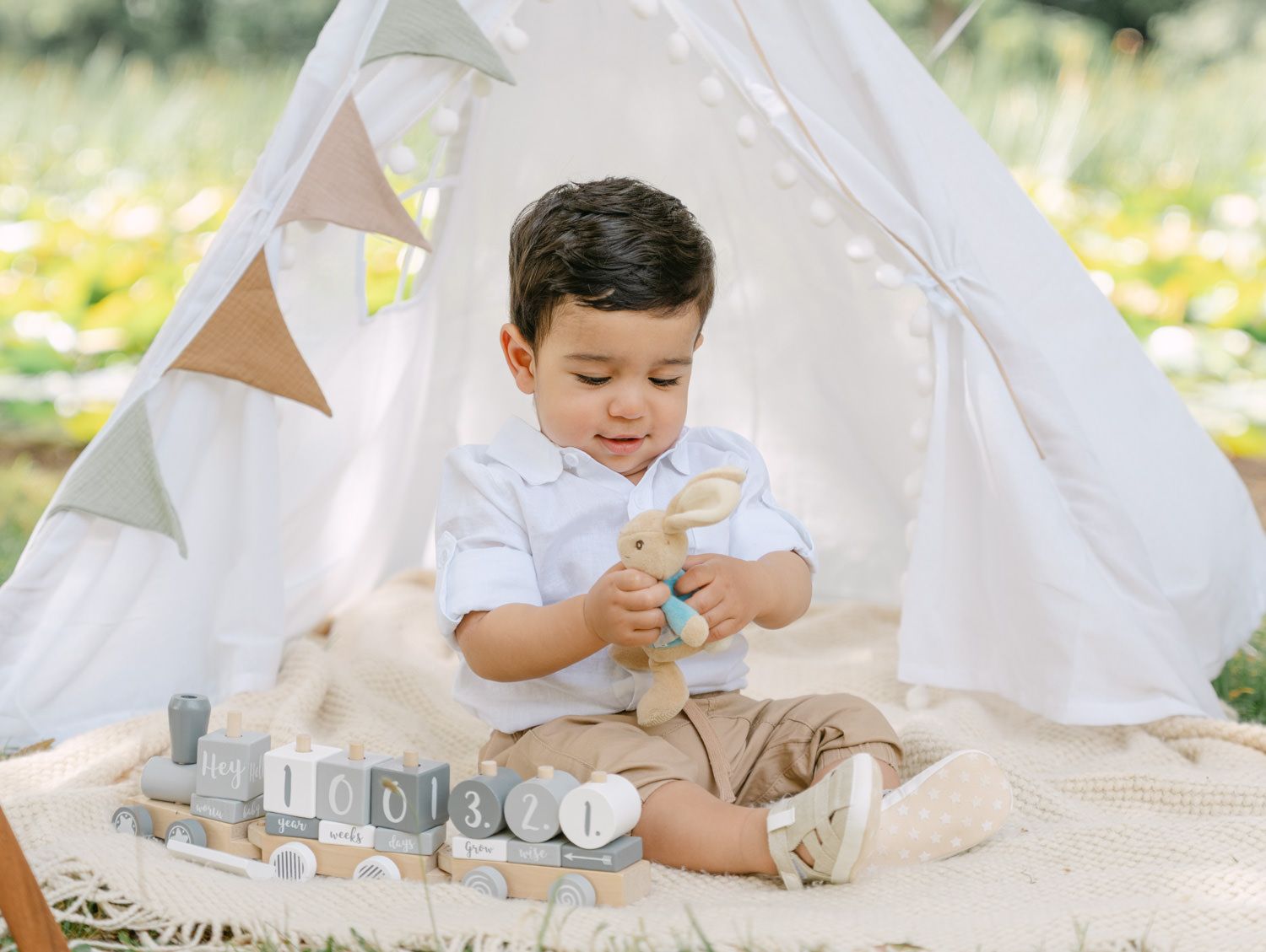 Toddler boy playing with wooden toys and bunny plush inside a white teepee tent outdoors.