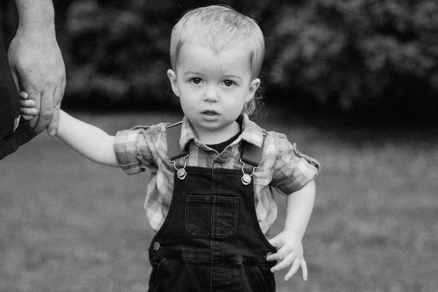 Black and white photo of a toddler boy in overalls holding an adults hand outdoors.