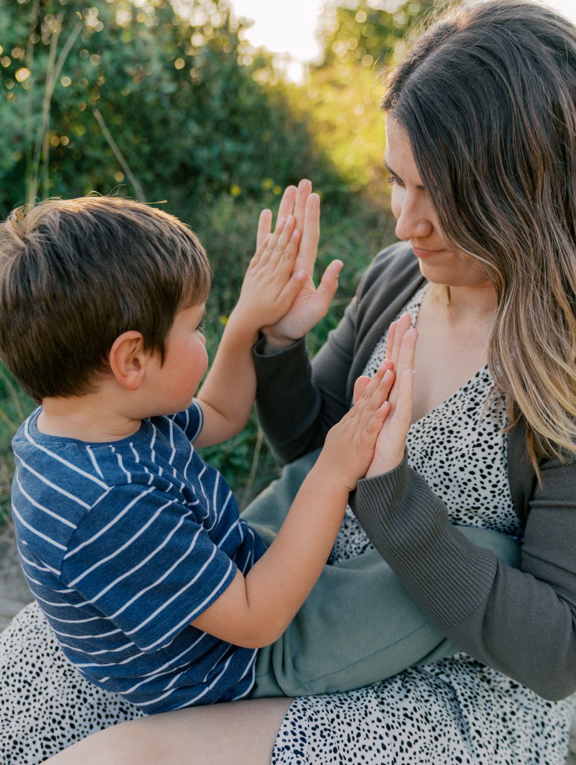 A mother and young boy share a joyful high-five moment outdoors surrounded by lush green trees.