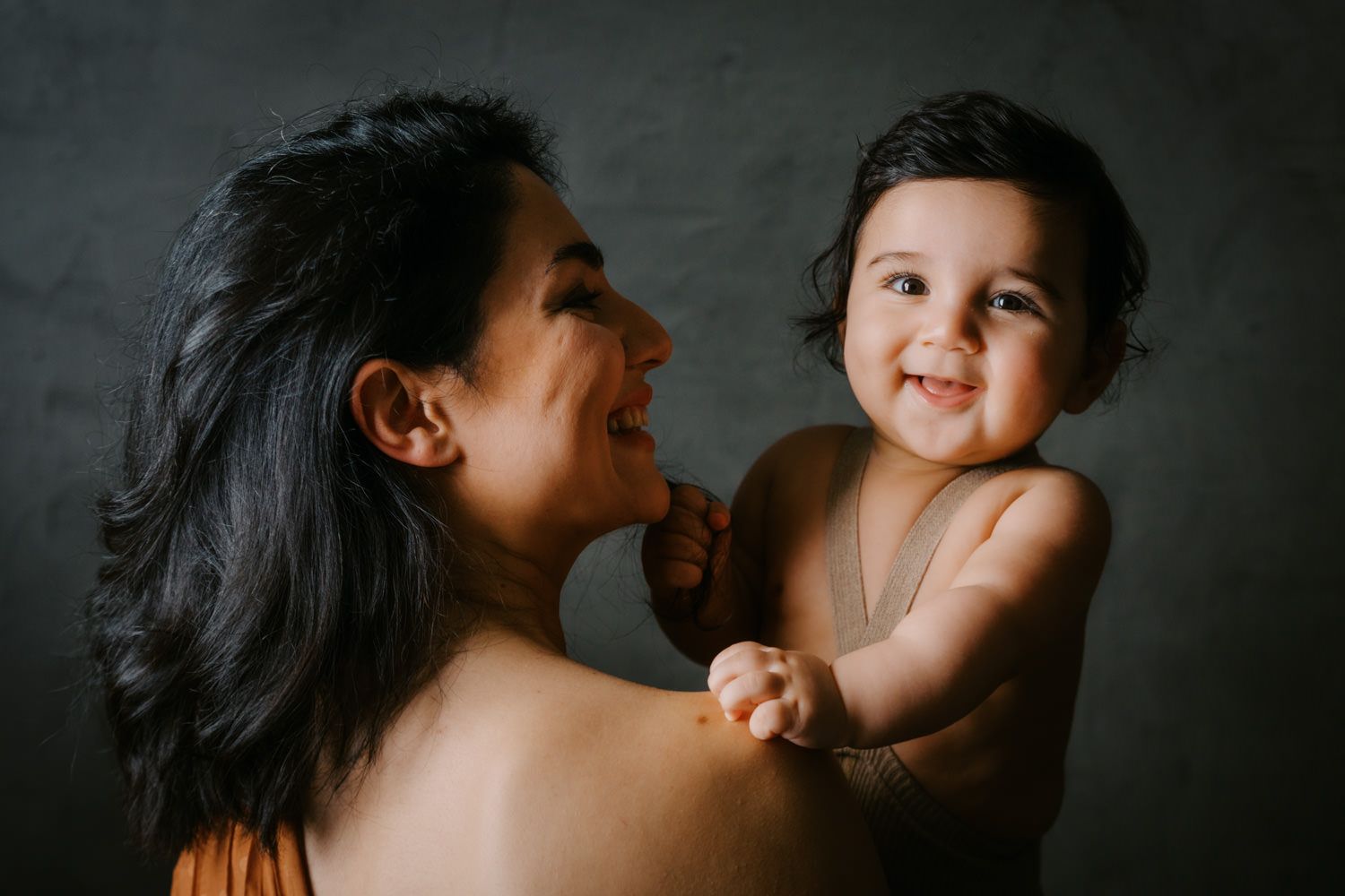 A smiling mother holds her happy, chubby baby against a dark gray studio background.