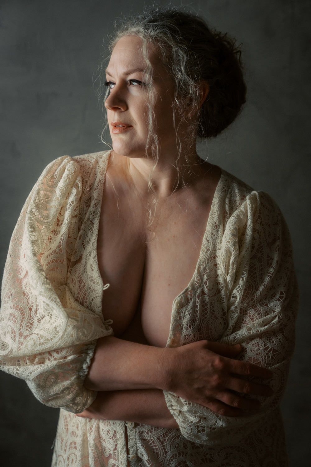 Mature woman in lace robe with arms crossed, looking sideways in dramatic studio portrait lighting.