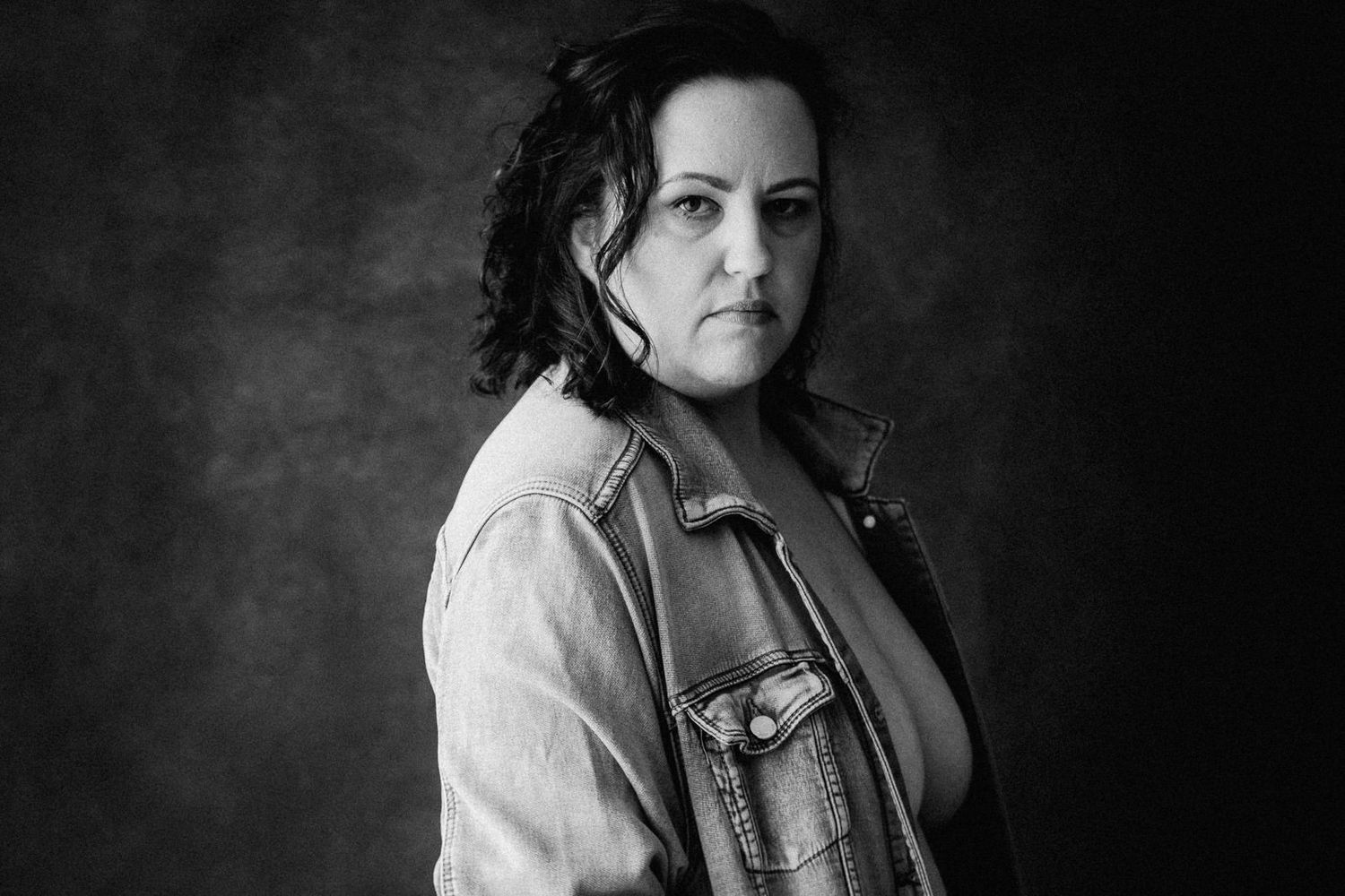 Black and white portrait of a woman in a denim jacket looking over her shoulder against a dark background.