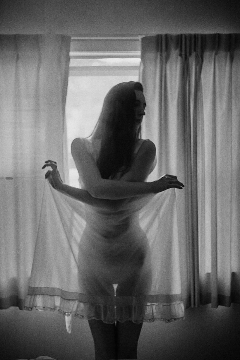 Black and white photo of a woman in a sheer white slip dress standing gracefully by a sunlit window.