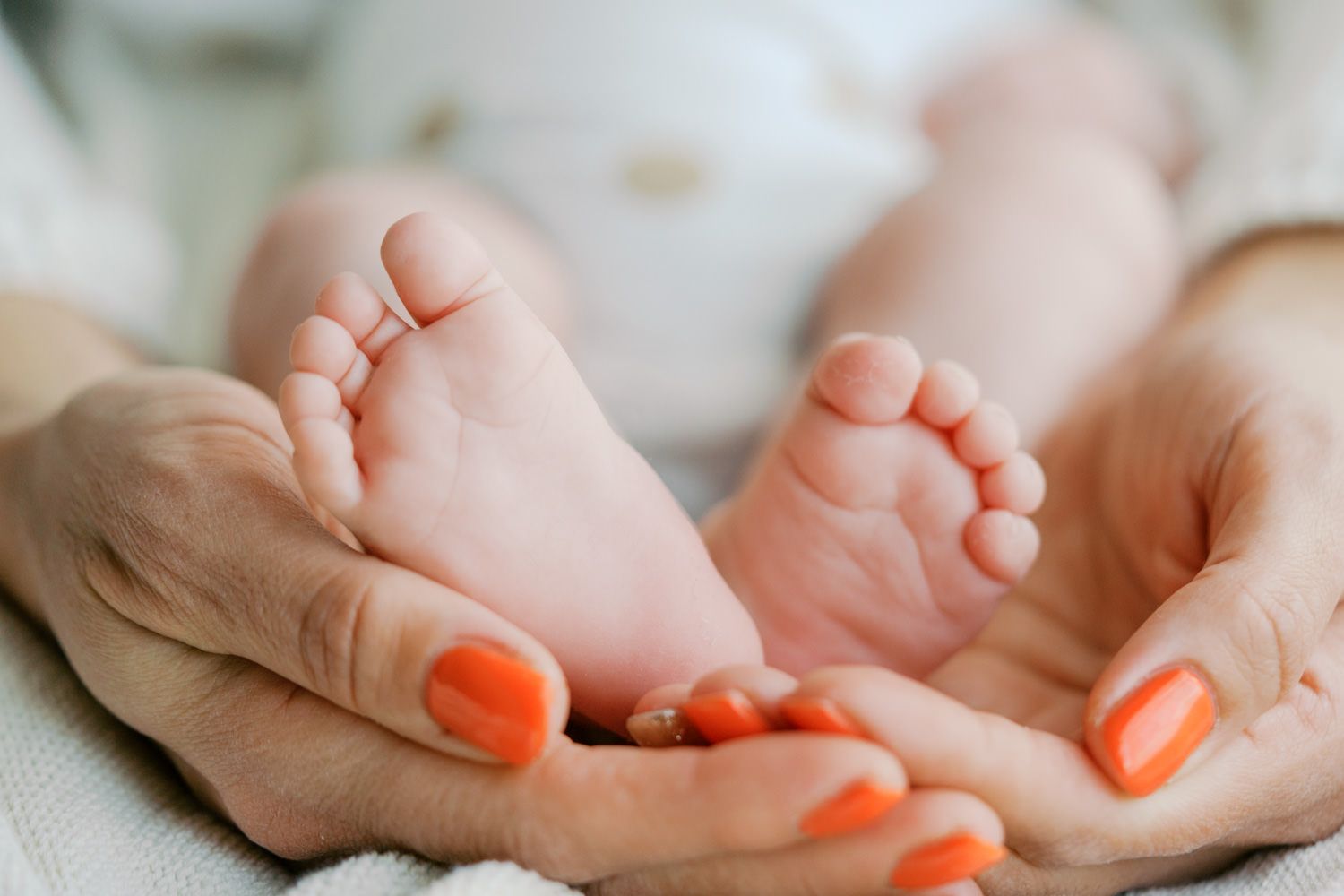 Close-up of a mother's hands with orange nails gently cradling tiny newborn baby feet.