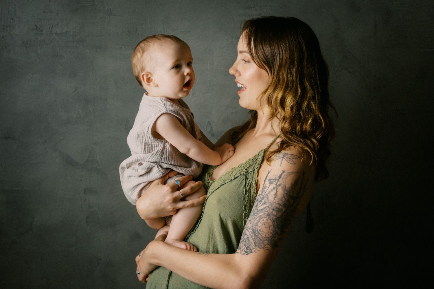 A tattooed mother in a green dress holds her baby in a studio portrait against a dark gray background.