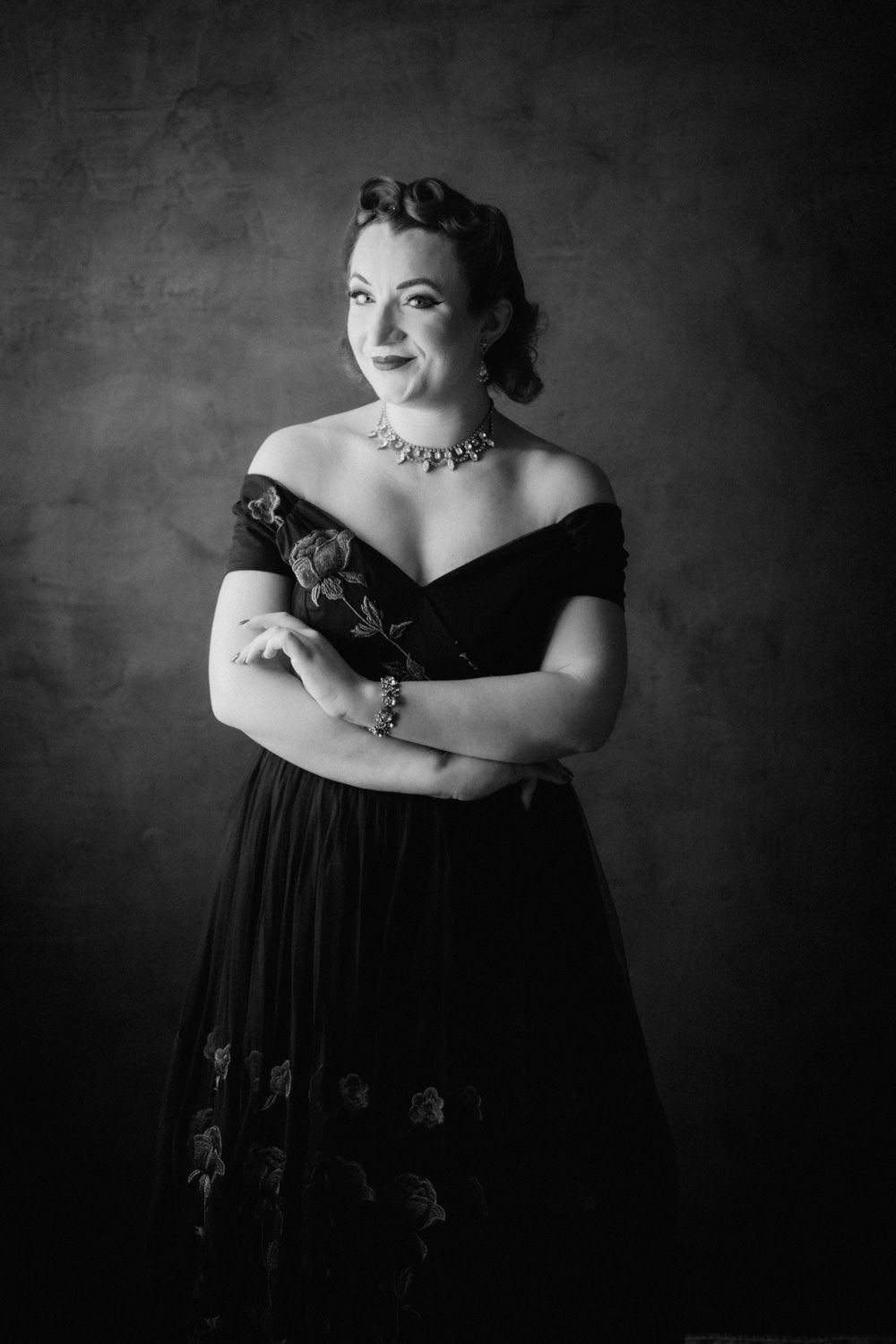 Black and white portrait of a woman in an off-shoulder gown with a necklace, arms crossed, posing elegantly.