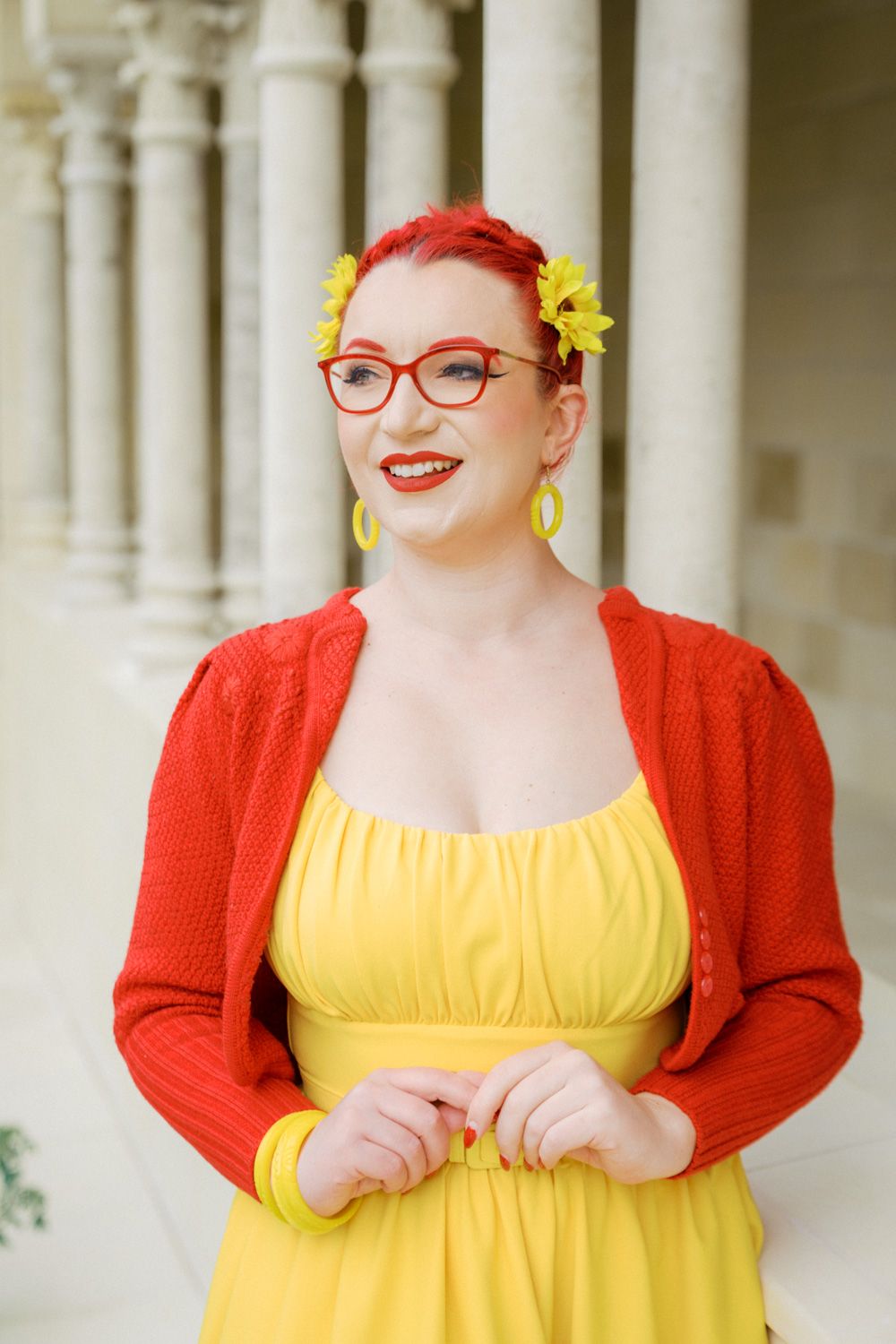Woman in yellow dress and red cardigan with red glasses and yellow flowers in her red hair smiling outdoors.
