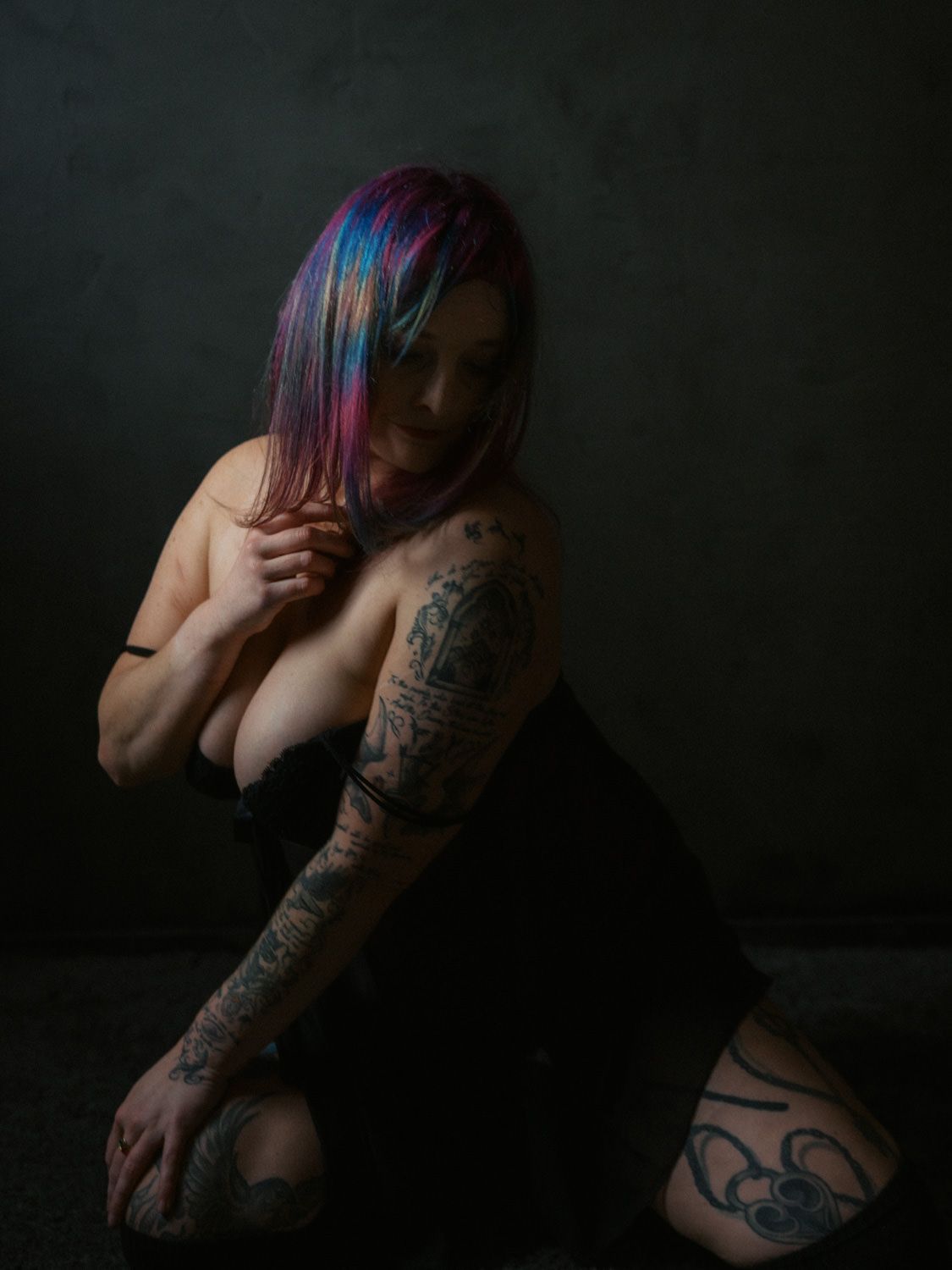 Woman with colorful pink and blue hair, covered in tattoos, wearing black, posed in dramatic dark studio lighting.
