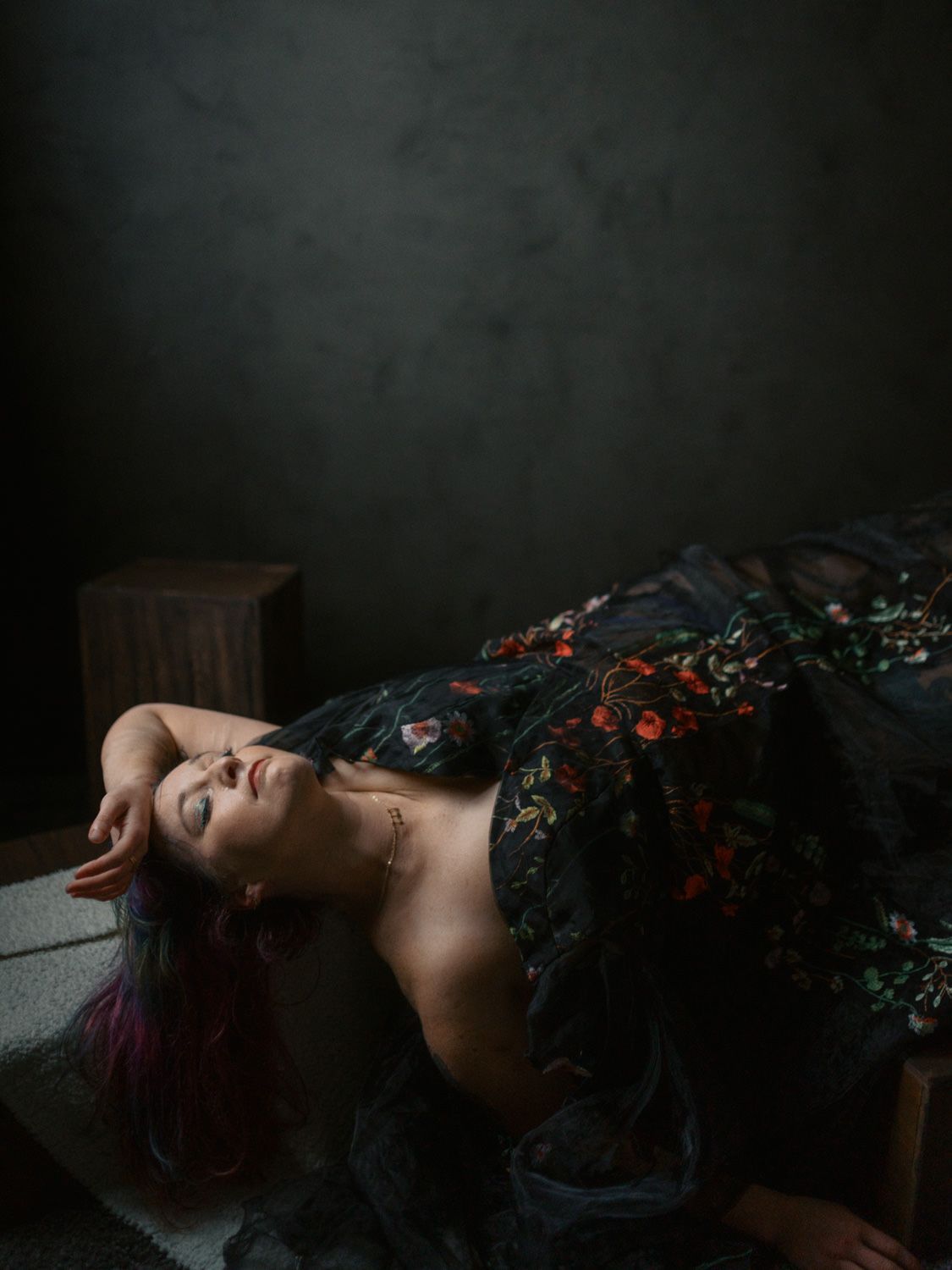 Woman in floral dark gown reclines dramatically against wooden furniture in moody studio portrait photography.