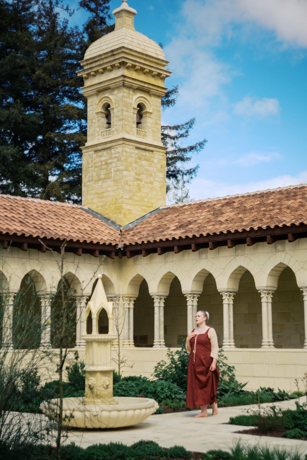 Woman in red dress stands in a sunlit Mediterranean-style courtyard with arched colonnade and stone bell tower.