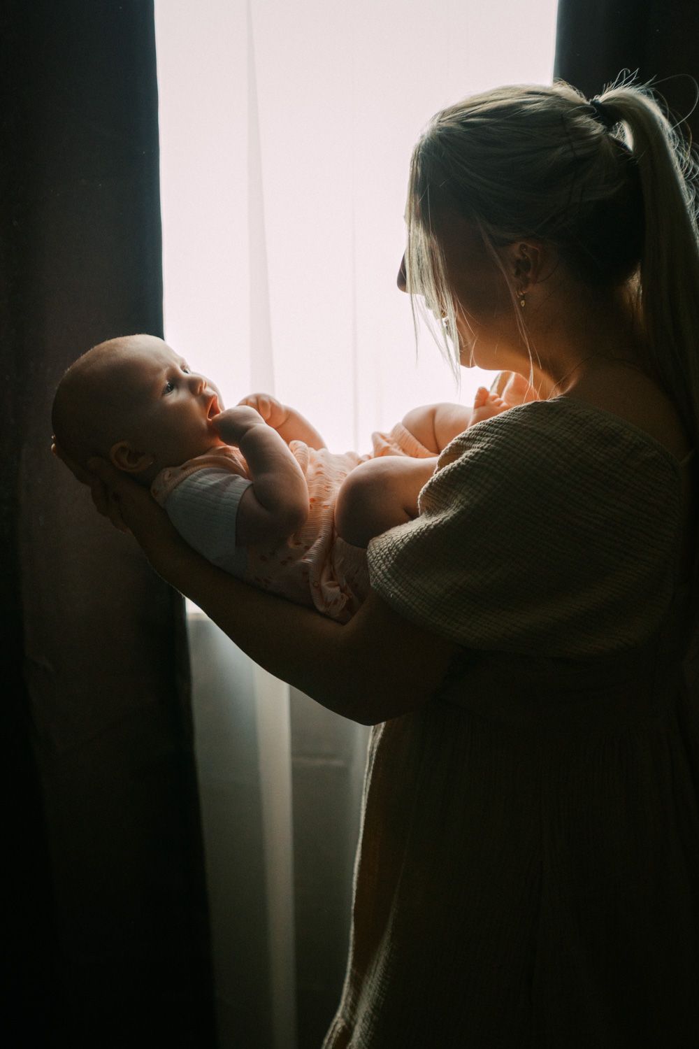 Mother holding newborn baby near bright window in soft natural light, tender moment of new parenthood.