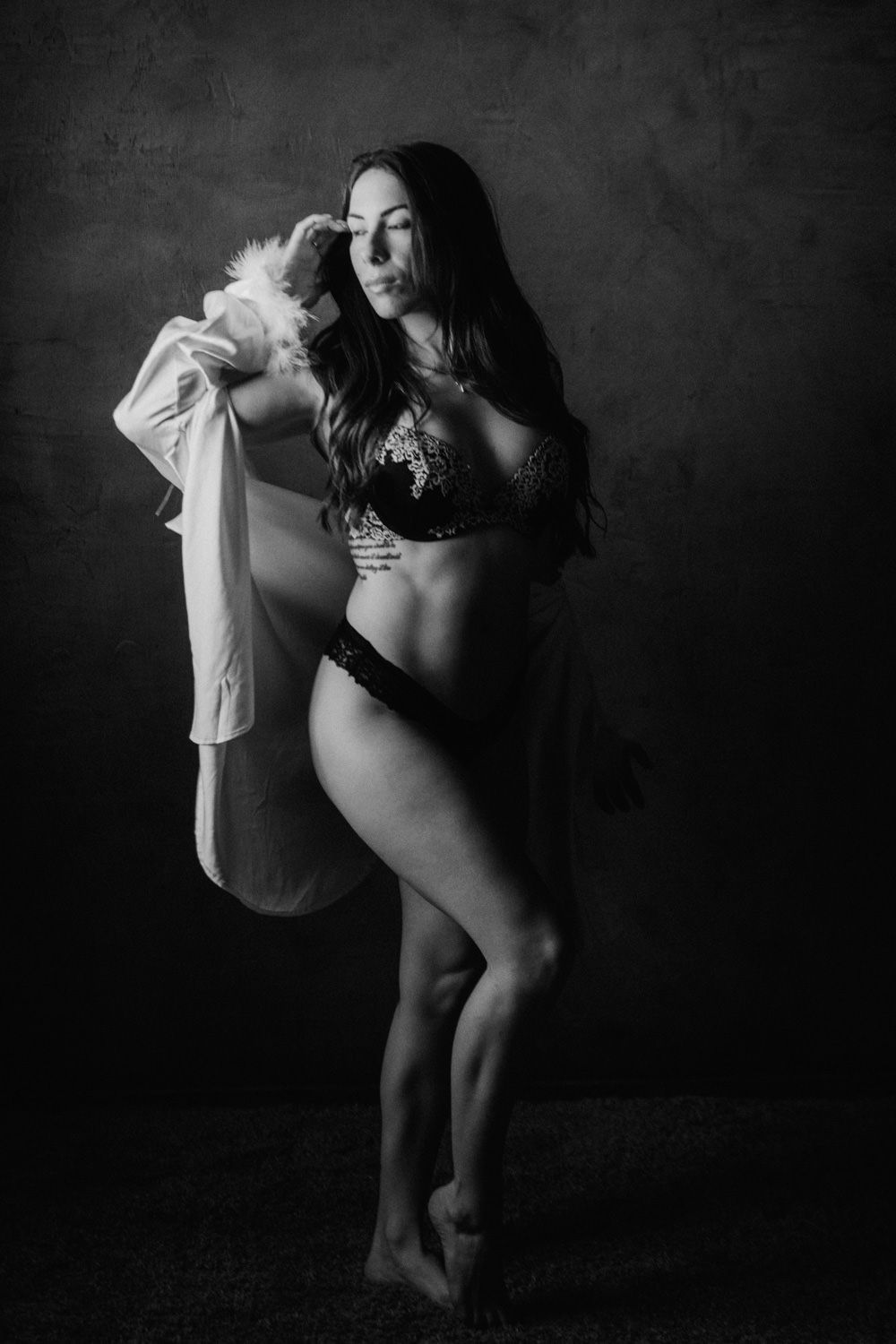 Black and white boudoir photo of a woman in lace lingerie holding an open white shirt in a studio.