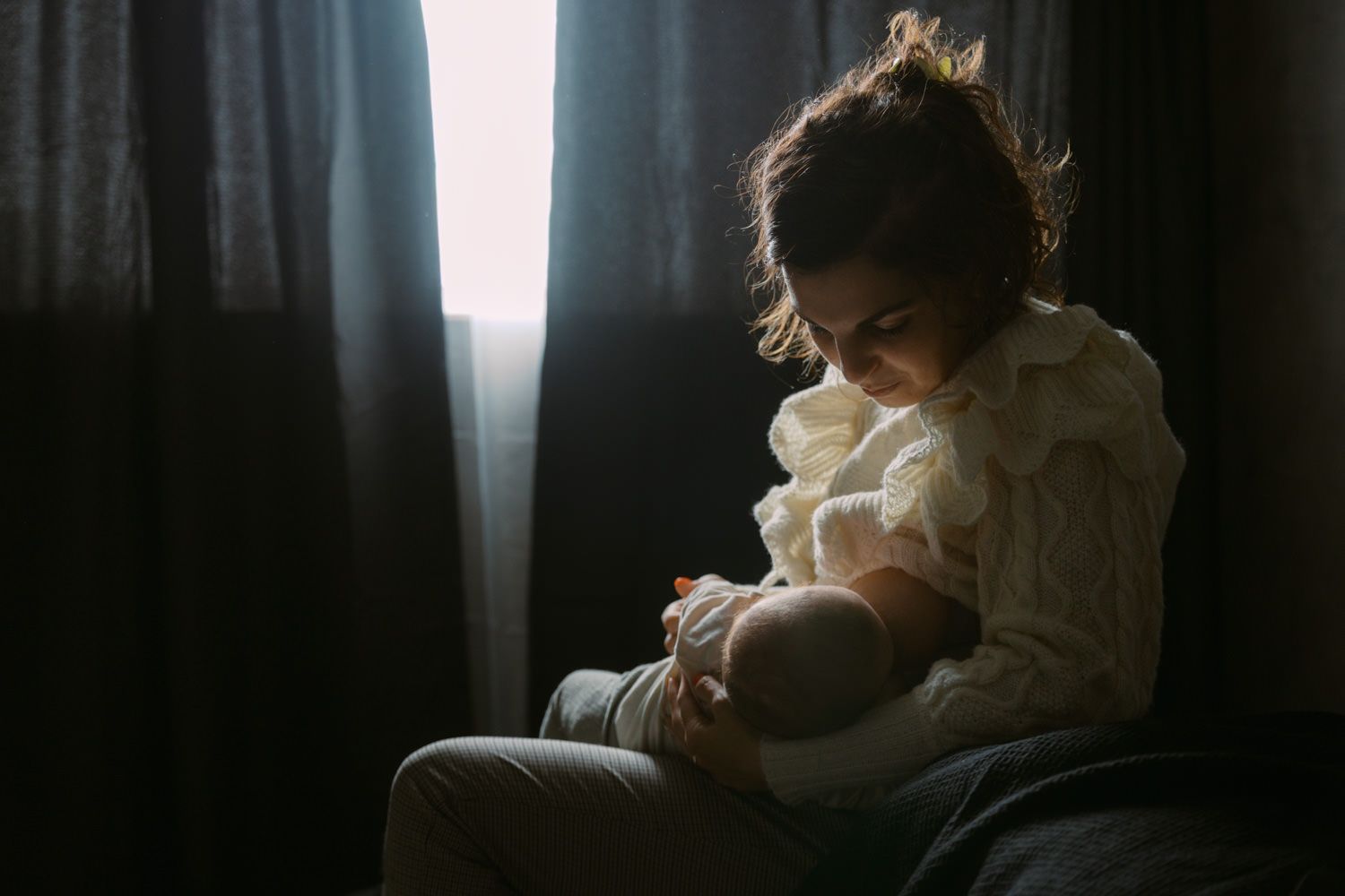 Young girl sitting by a dark window, cradling a baby doll in soft, moody light.