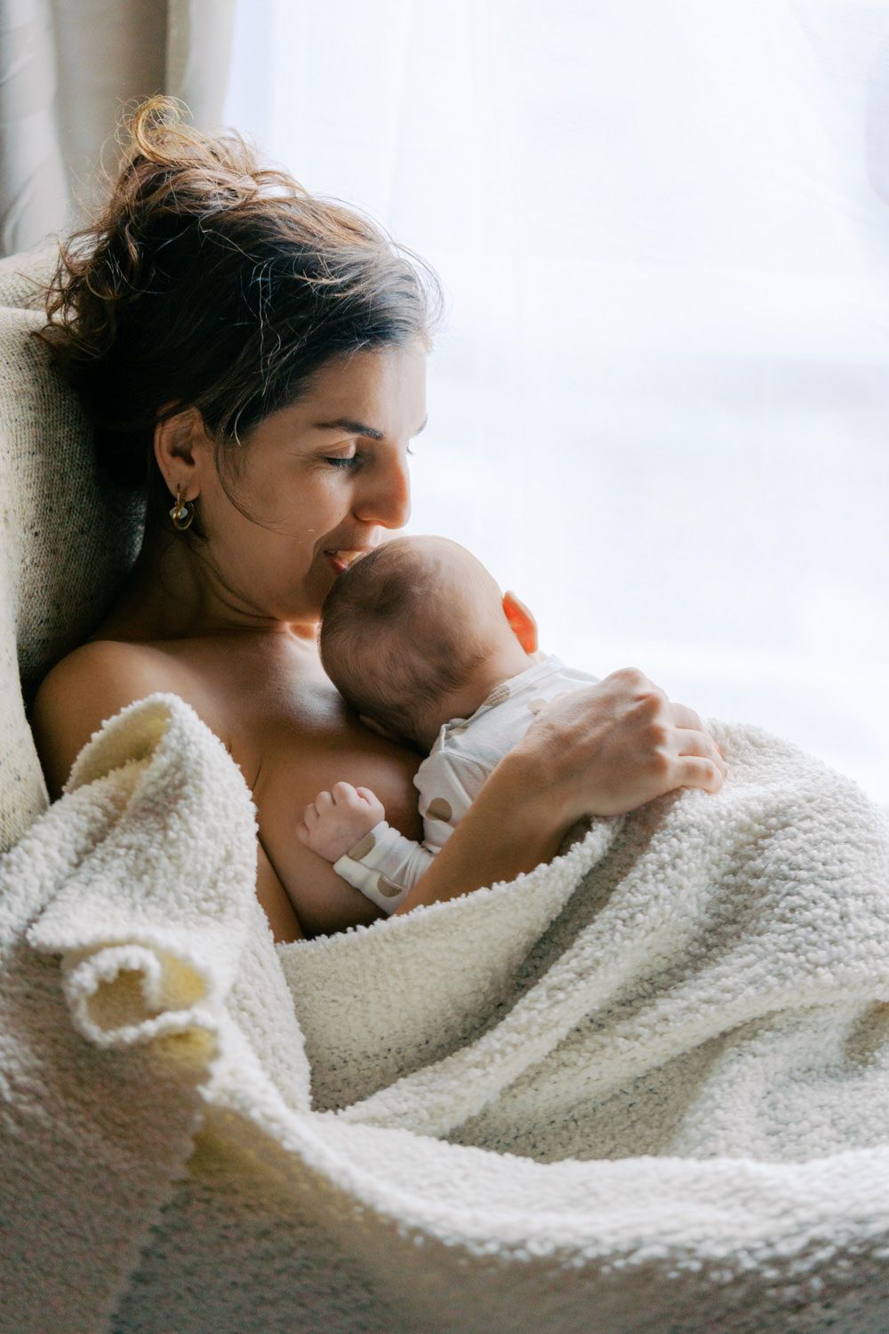 A tender moment of a mother cradling her newborn baby wrapped in a soft white blanket by a bright window.