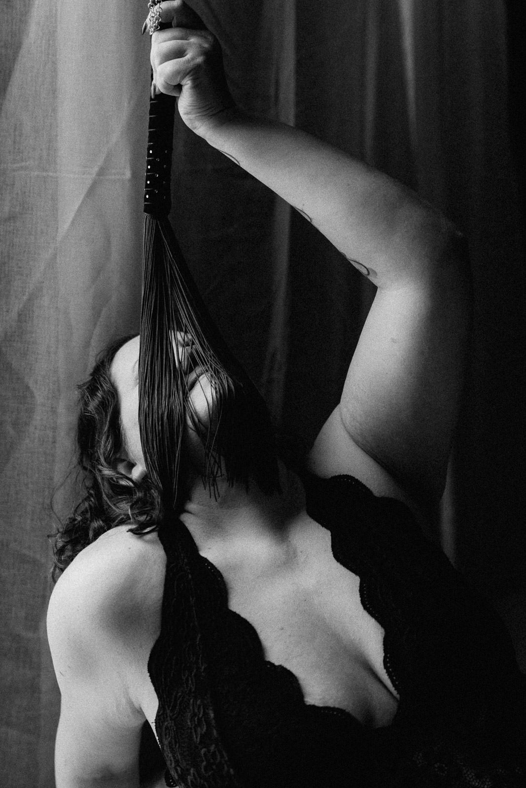 Black and white boudoir photo of a woman in lace lingerie holding a flogger whip against a draped backdrop.