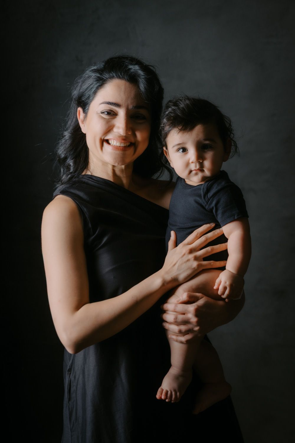 Smiling woman in black dress holding a baby in a black onesie against a dark studio background.