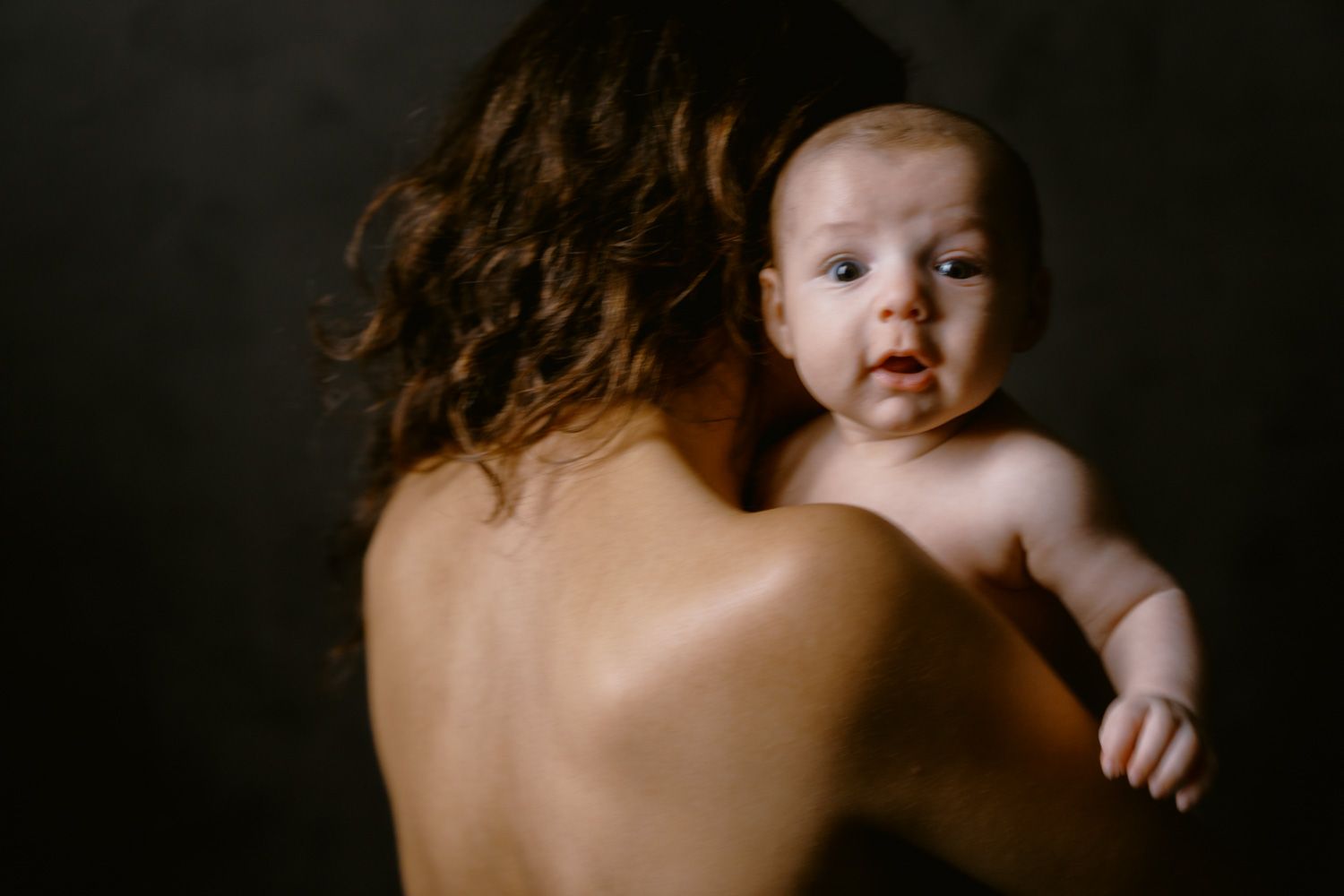 A mother holds her newborn baby against her bare shoulder in a tender, intimate portrait with dark background.