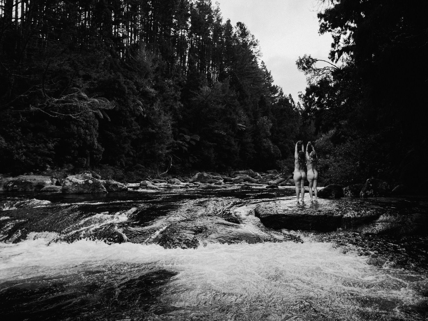 Black and white photo of two people standing on rocks beside a rushing river surrounded by dense forest.