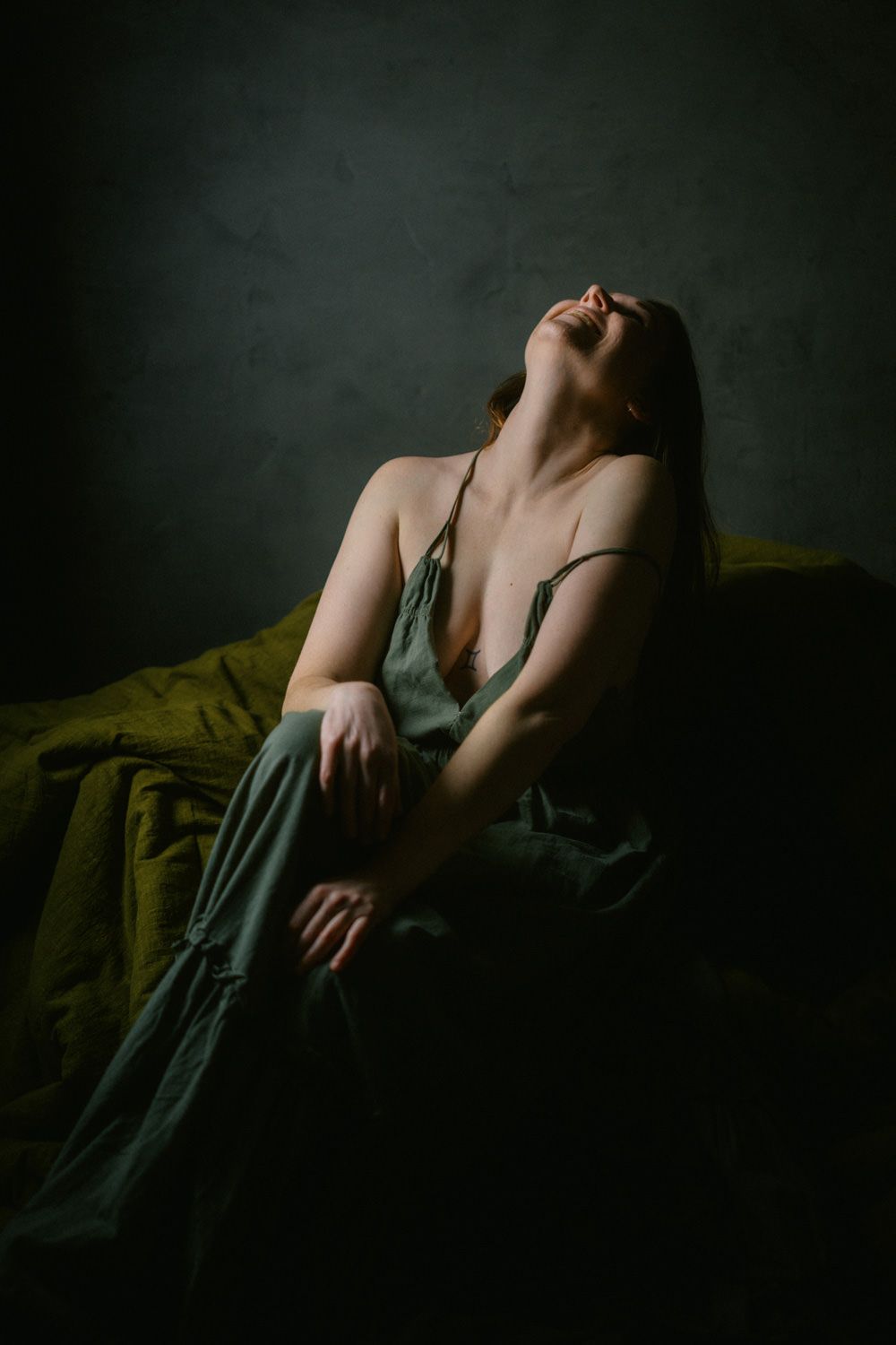 A woman in a green slip dress sits on a bed with olive bedding, head tilted back, in moody dramatic lighting.