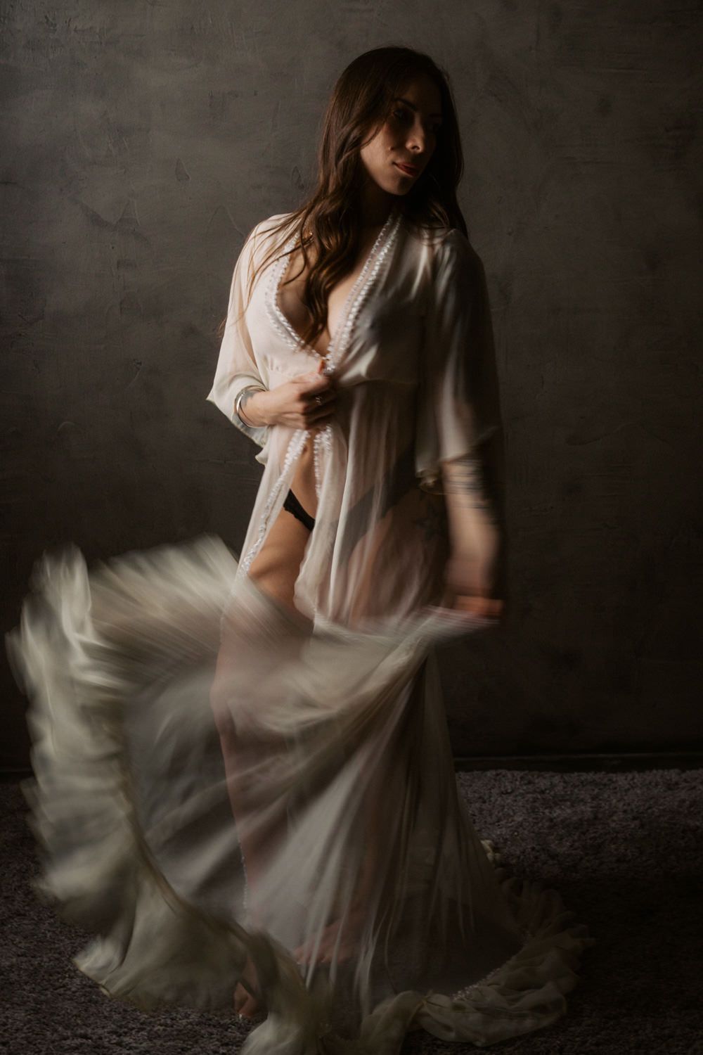 A woman in a flowing white sheer robe poses dramatically in a dimly lit studio, her skirt captured in motion blur.