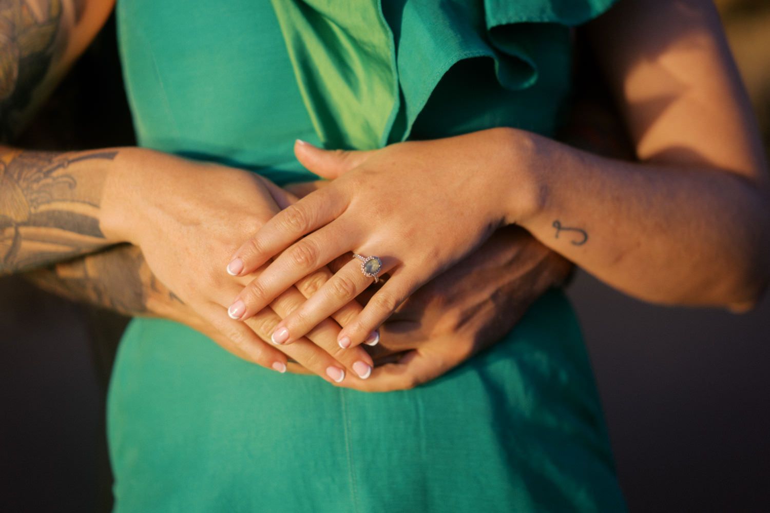 Close-up of couple holding hands showing engagement ring, woman in teal dress with tattoos visible.