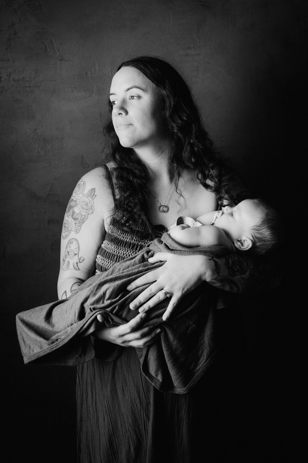 Black and white portrait of a tattooed mother breastfeeding her newborn baby while gazing thoughtfully to the side.
