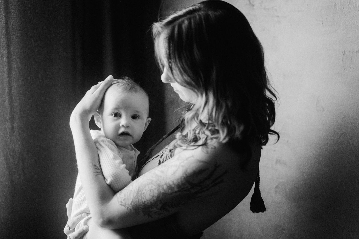 Black and white photo of a tattooed mother tenderly holding her baby near a window.