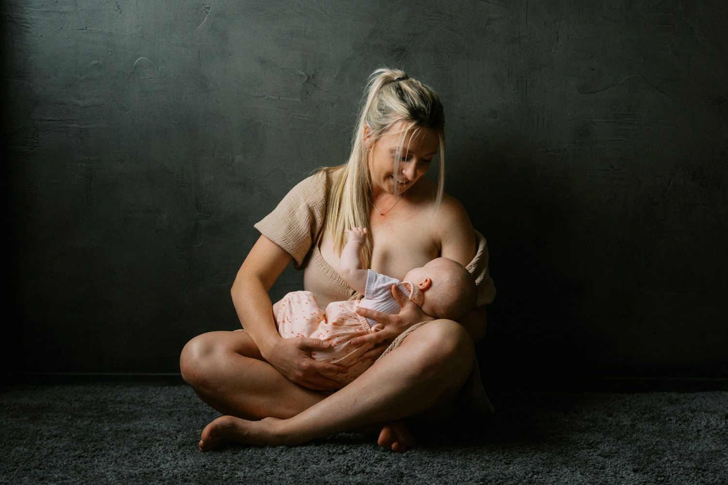 Mother breastfeeding newborn baby while sitting cross-legged on floor against dark gray wall.