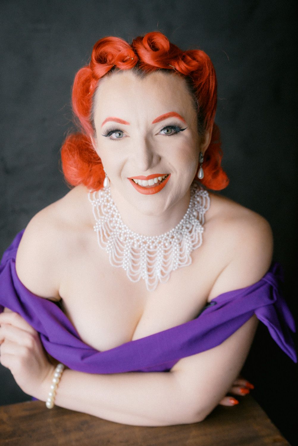 Vintage pin-up style woman with red hair, pearl necklace, and purple off-shoulder wrap smiling at camera.