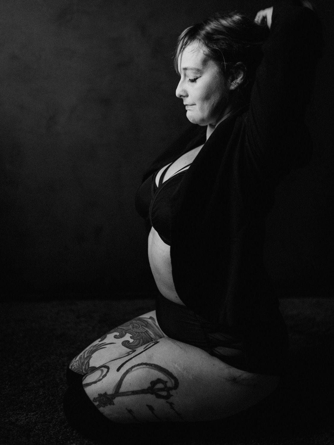 Black and white photo of a pregnant woman with tattoos sitting peacefully with eyes closed in a serene pose.