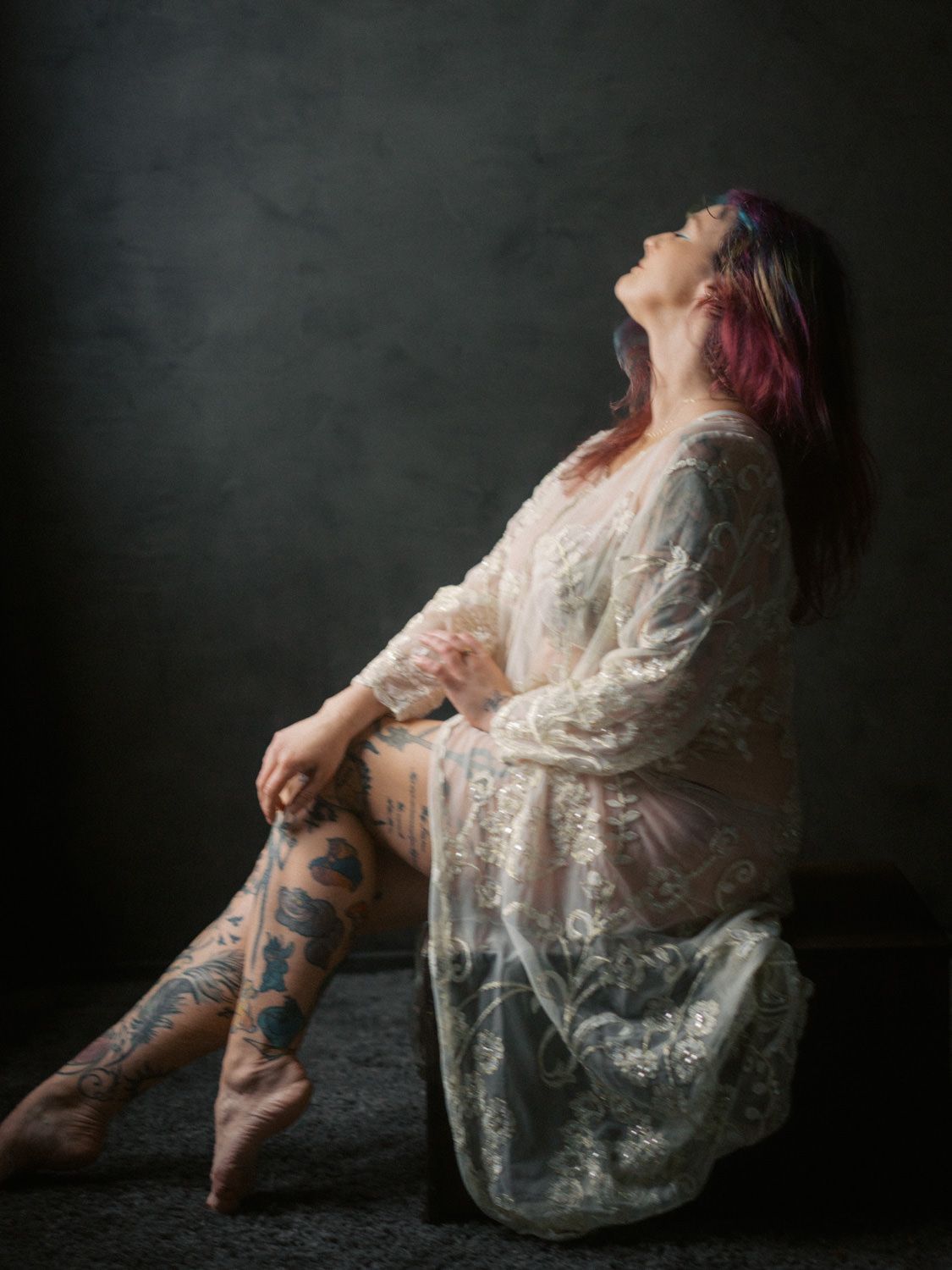 Woman with pink hair and tattoos poses in white lace robe against dark backdrop.