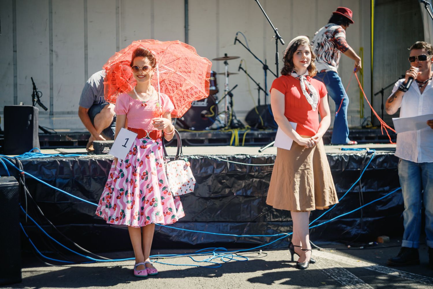 Two women in vintage 1950s outfits stand near a stage at an outdoor festival, one holding a red parasol.