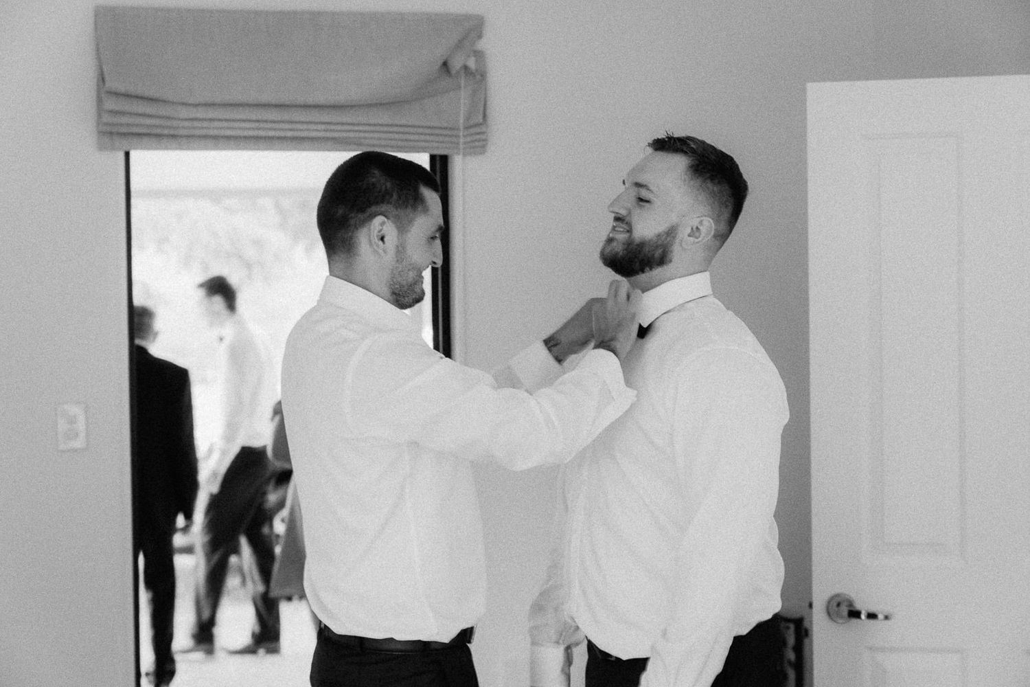 Two men in white dress shirts help each other with ties while getting ready for a wedding ceremony.
